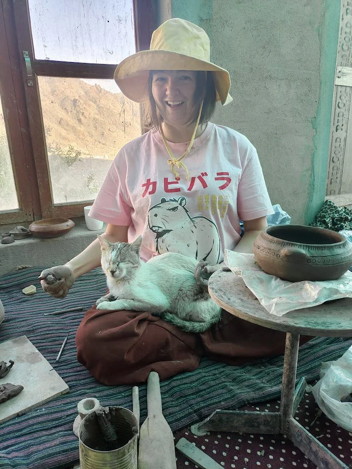A woman wearing a wide-brimmed hat and a pink T-shirt with a lion graphic is sitting cross-legged on a cloth-covered surface indoors. She is holding a gray and white cat on her lap, and they are near pottery and sculpting tools. Behind her is a windo