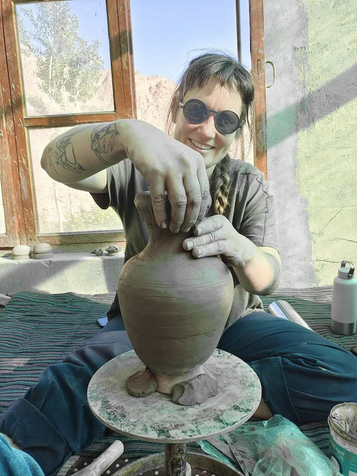 A woman with sunglasses and a braid is shaping a large clay pot on a pottery wheel. She is smiling and working indoors near a window with a scenic outdoor view.
