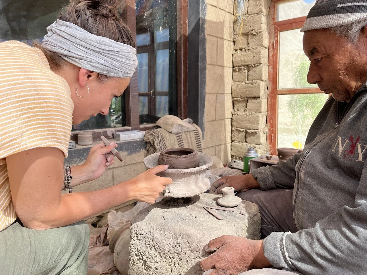 A woman and a man working on pottery inside a workshop with brick walls and a window. The woman is shaping a clay pot, while the man is observing.