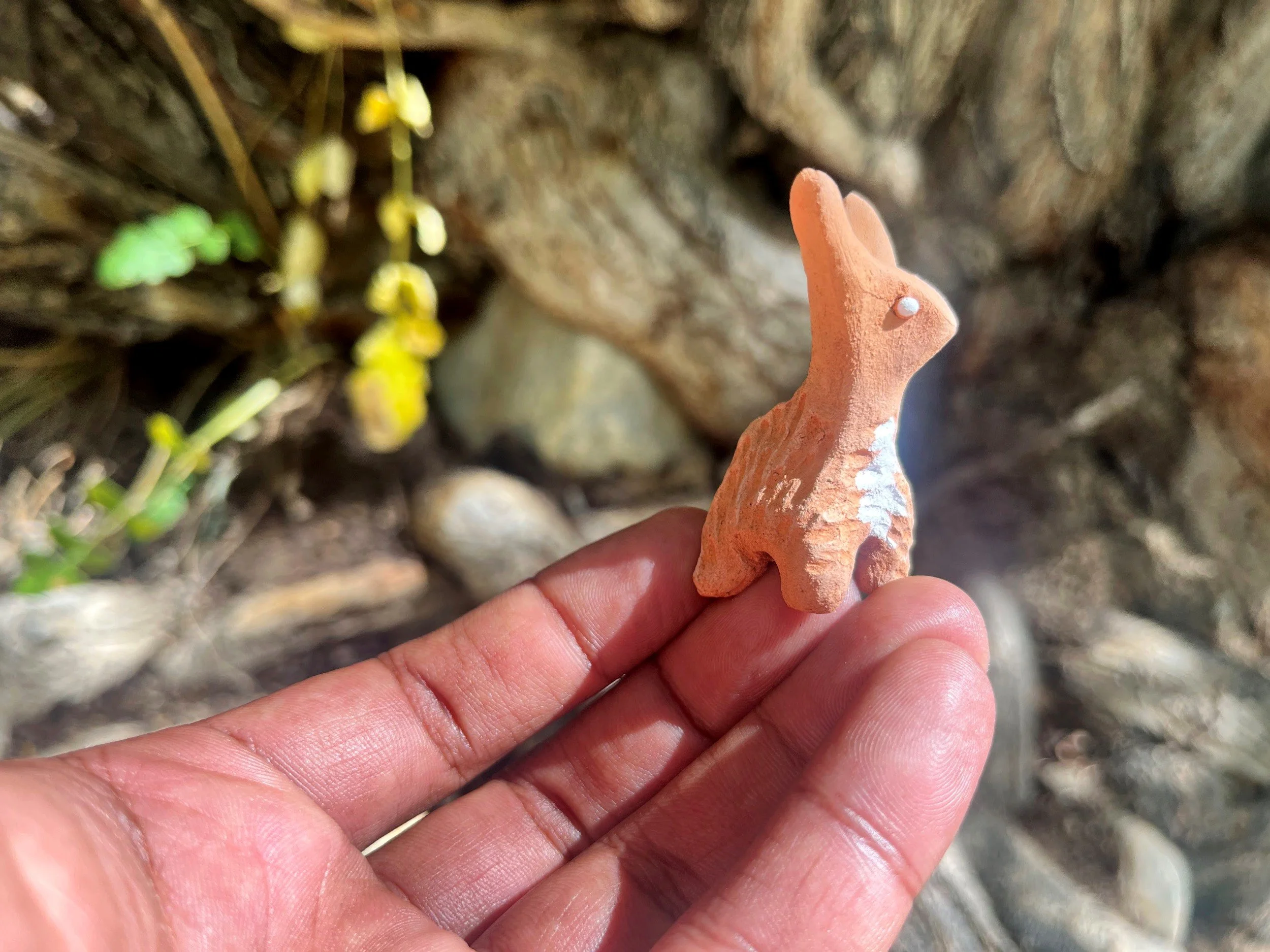 A person holding a small, orange, textured figurine or sculpture of an animal, possibly a yak, with a background of rocks and plants.