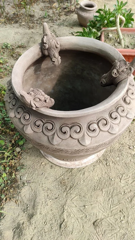 Decorative round stone planter with dragon and lion head sculptures around the rim, situated outdoors on dirt ground with plants and pottery nearby.