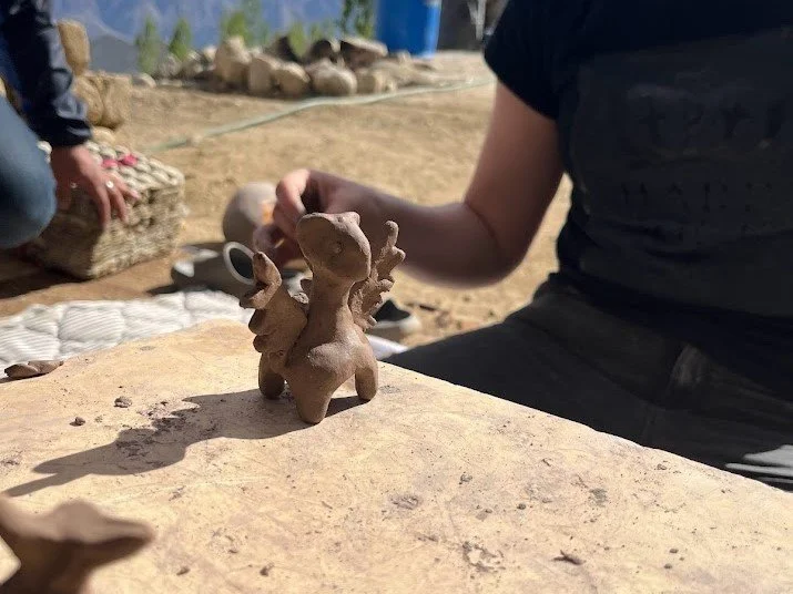Person in black shirt working with clay to create a small sculpture of a mouse with big ears, in an outdoor studio.