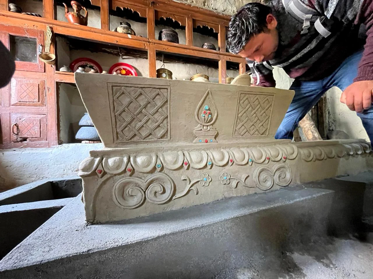 A man inspecting a handcrafted clay stove (thap) with intricate carvings and colorful inlay details, inside a rustic room with wooden shelves and traditional kitchenware.