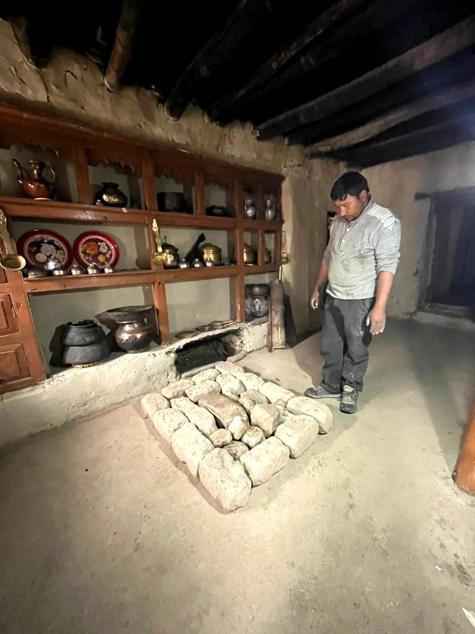 A young man looking down at a small rectangular arrangement of rocks inside a rustic room with clay walls and wooden ceiling beams. There is a wooden shelf with various metal and ceramic containers in the background.