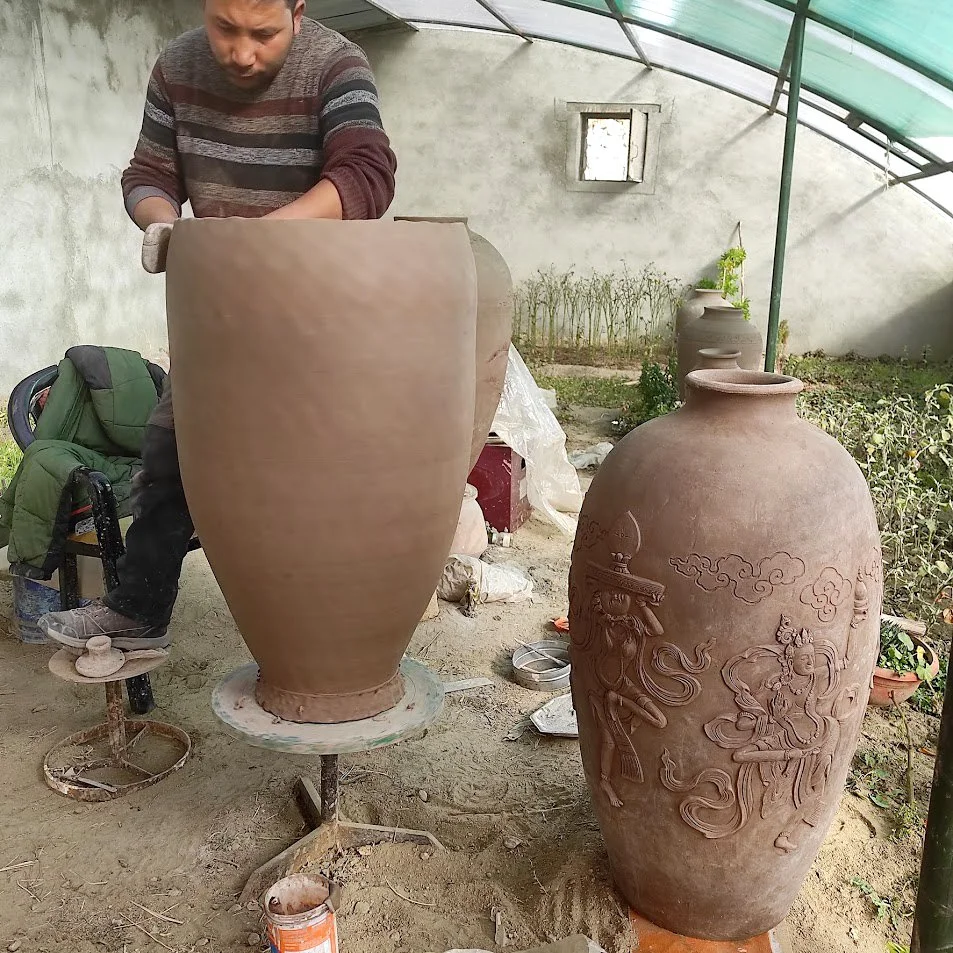 A man working on shaping a large, unfinished clay vase, with another decorated clay vase in the background, in a pottery studio or outdoor workshop.