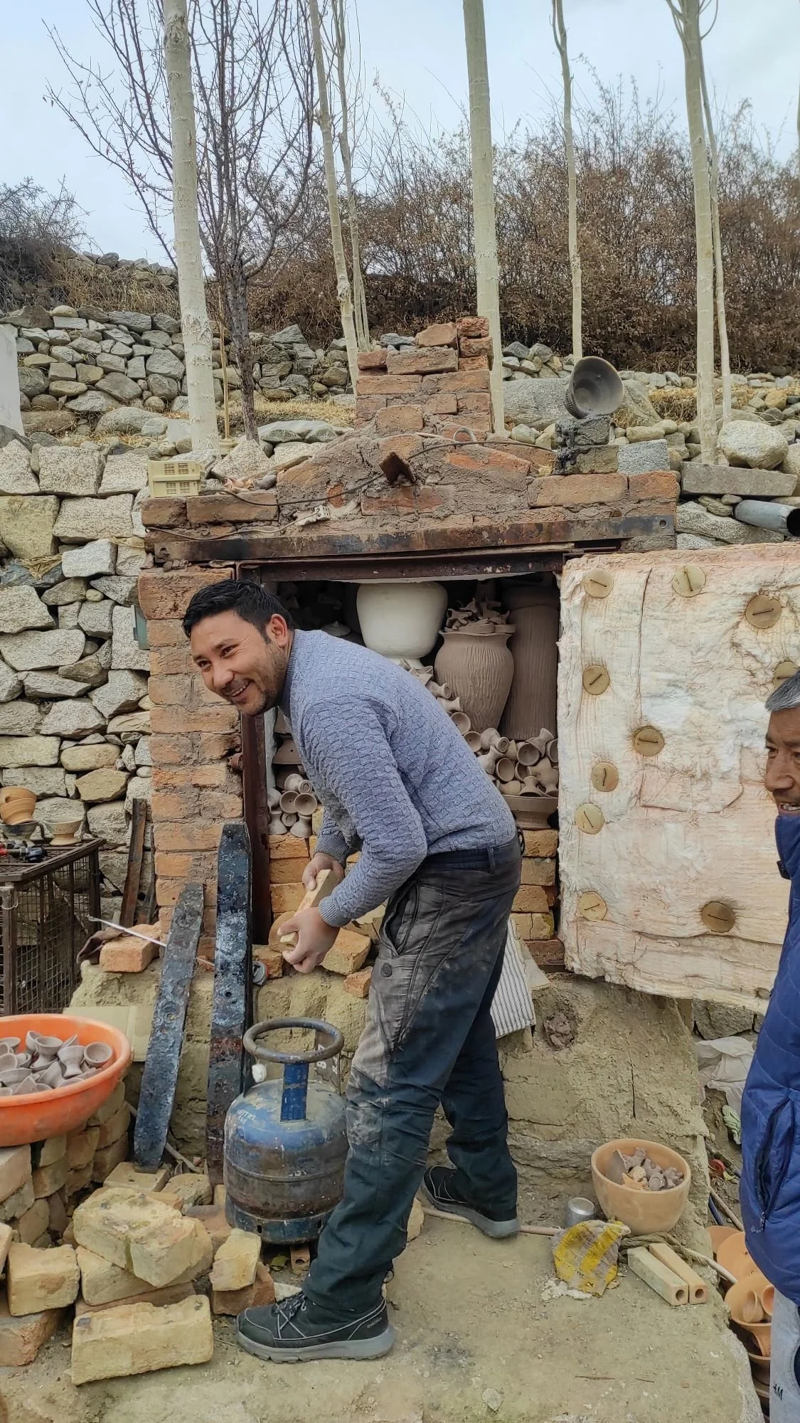 Men working on a brick and pottery kiln outdoors with trees and rocks in the background.