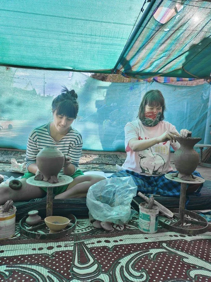 Two women engaged in pottery making inside a tent, sitting cross-legged with pottery wheels and tools around them.