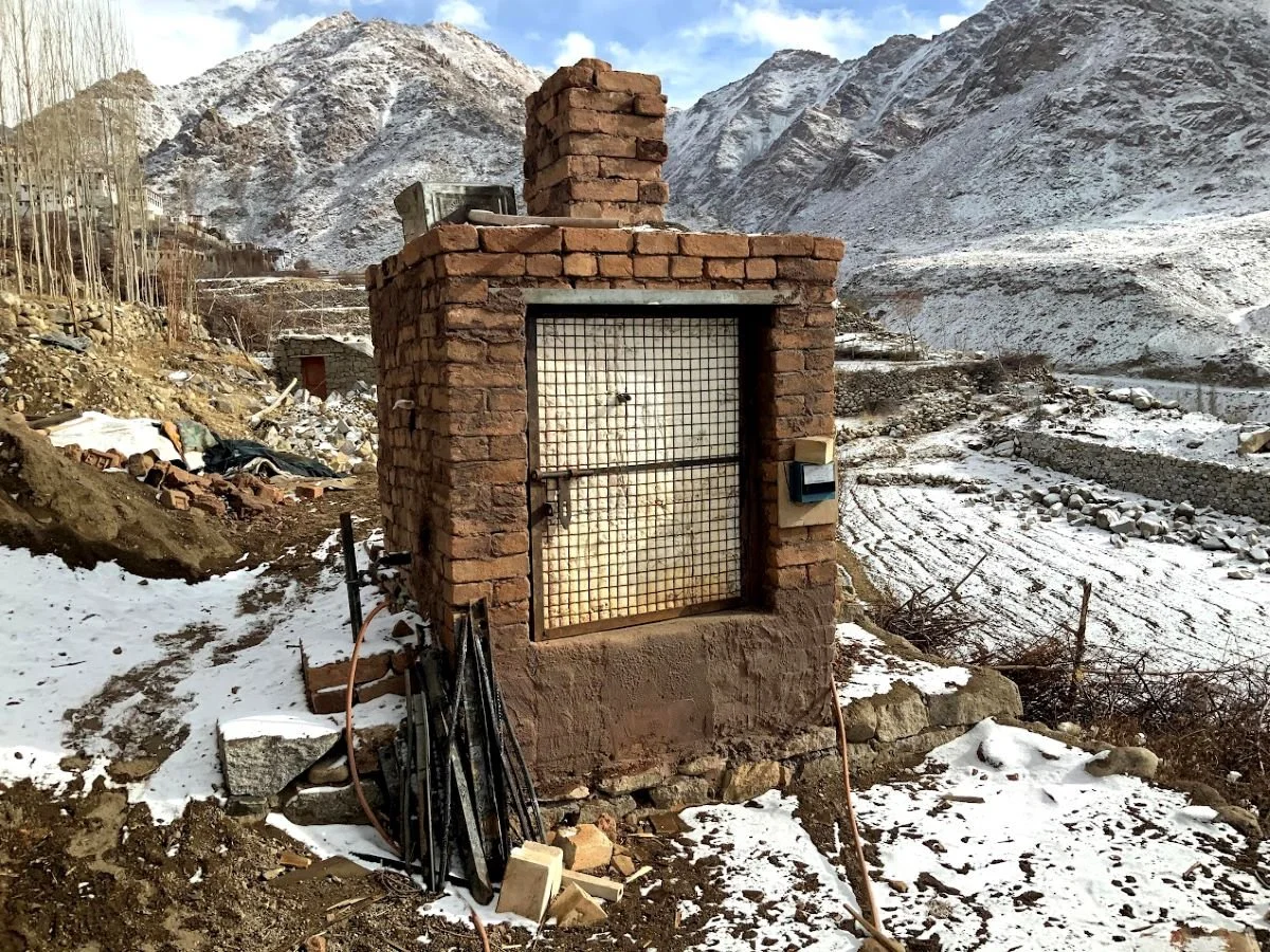 A small brick kiln with a gate and a lock, situated in a snowy mountainous landscape. There are tools and wires leaning against it, and mountains with snow in the background.