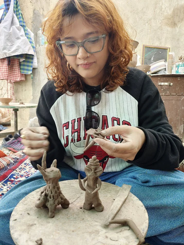 A woman with glasses and curly hair sculpting small animal figures from clay on a wheel in an outdoor workshop.