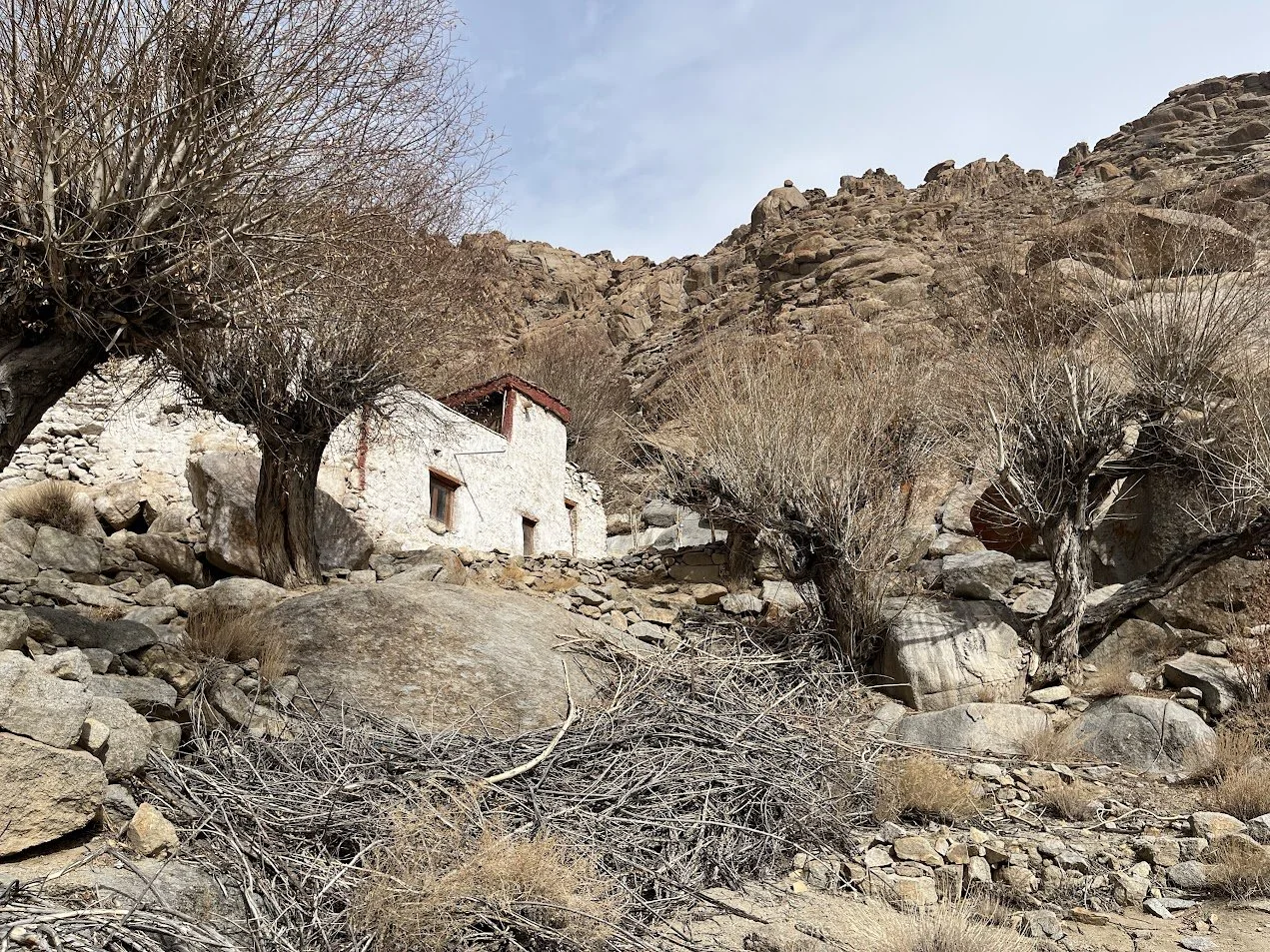 A small white house with a red roof situated on a rocky slope among leafless trees in a mountainous desert landscape.