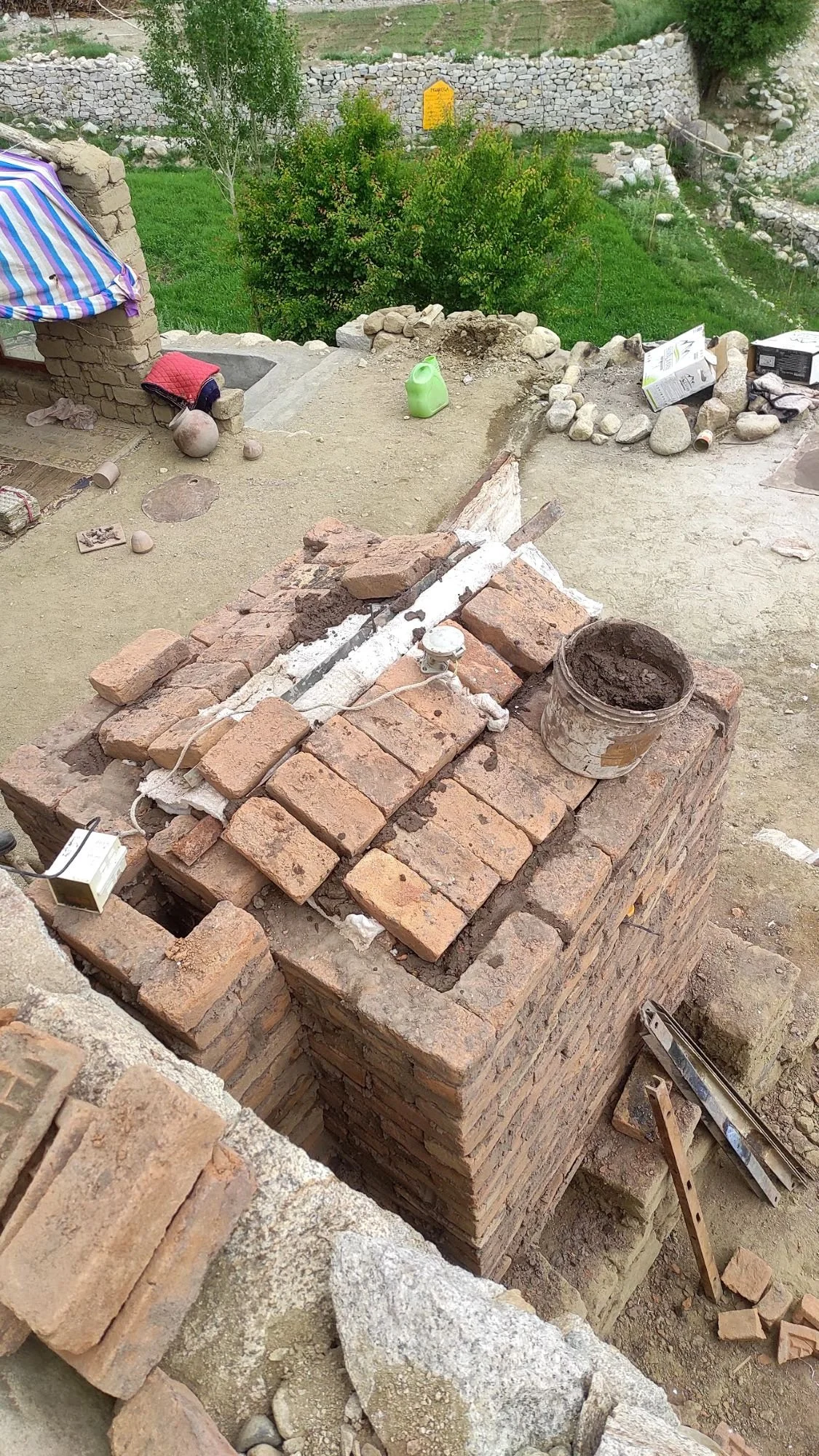A brick kiln under construction with a bucket of mortar on top. Various construction tools and materials are scattered around, with a garden and trees in the background.