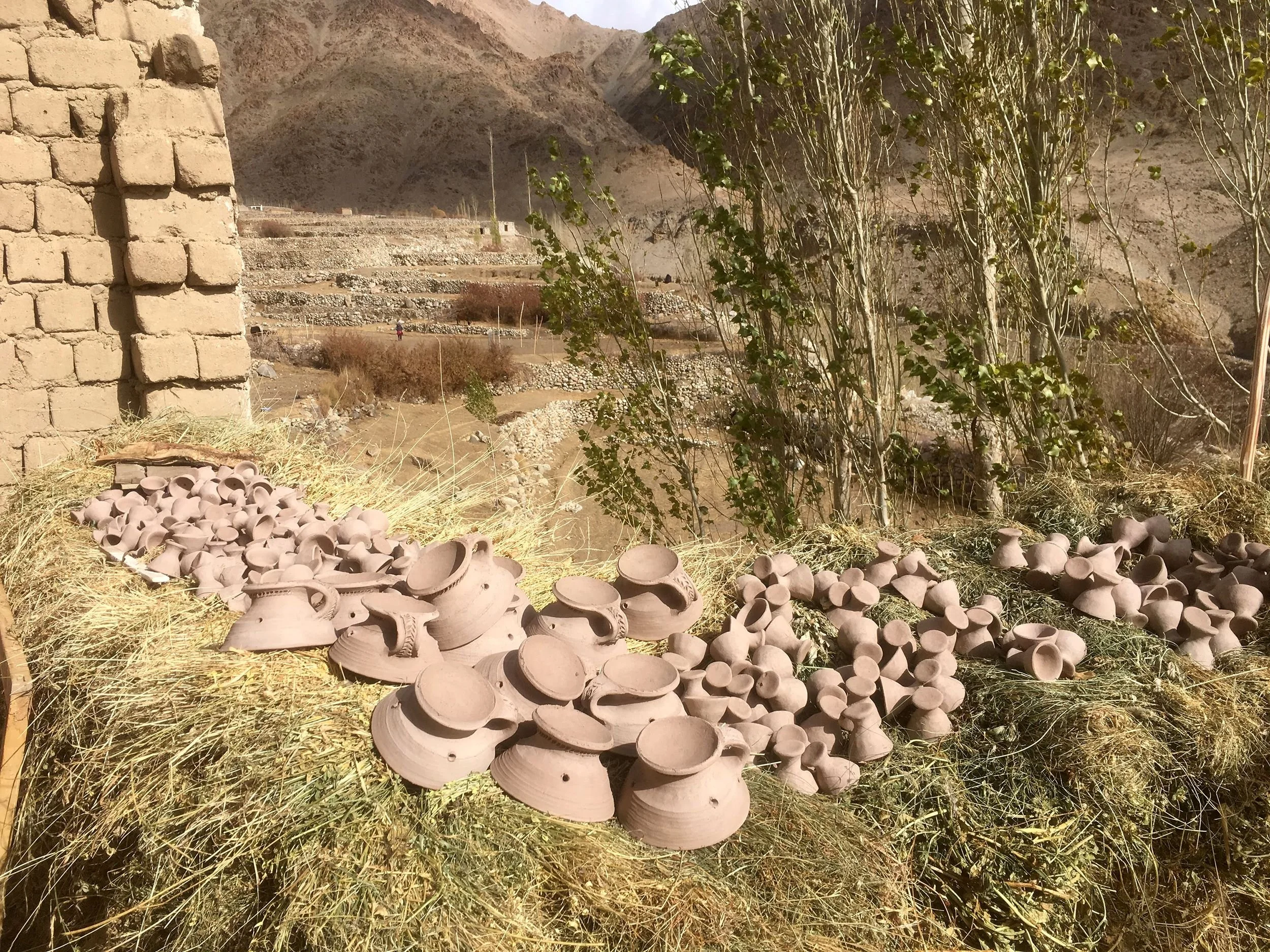 A landscape of terraced agricultural fields on a hillside with rocky mountains in the background. In the foreground, there are many unfinished clay pots scattered for drying on yellow straw and some trees with sparse foliage.