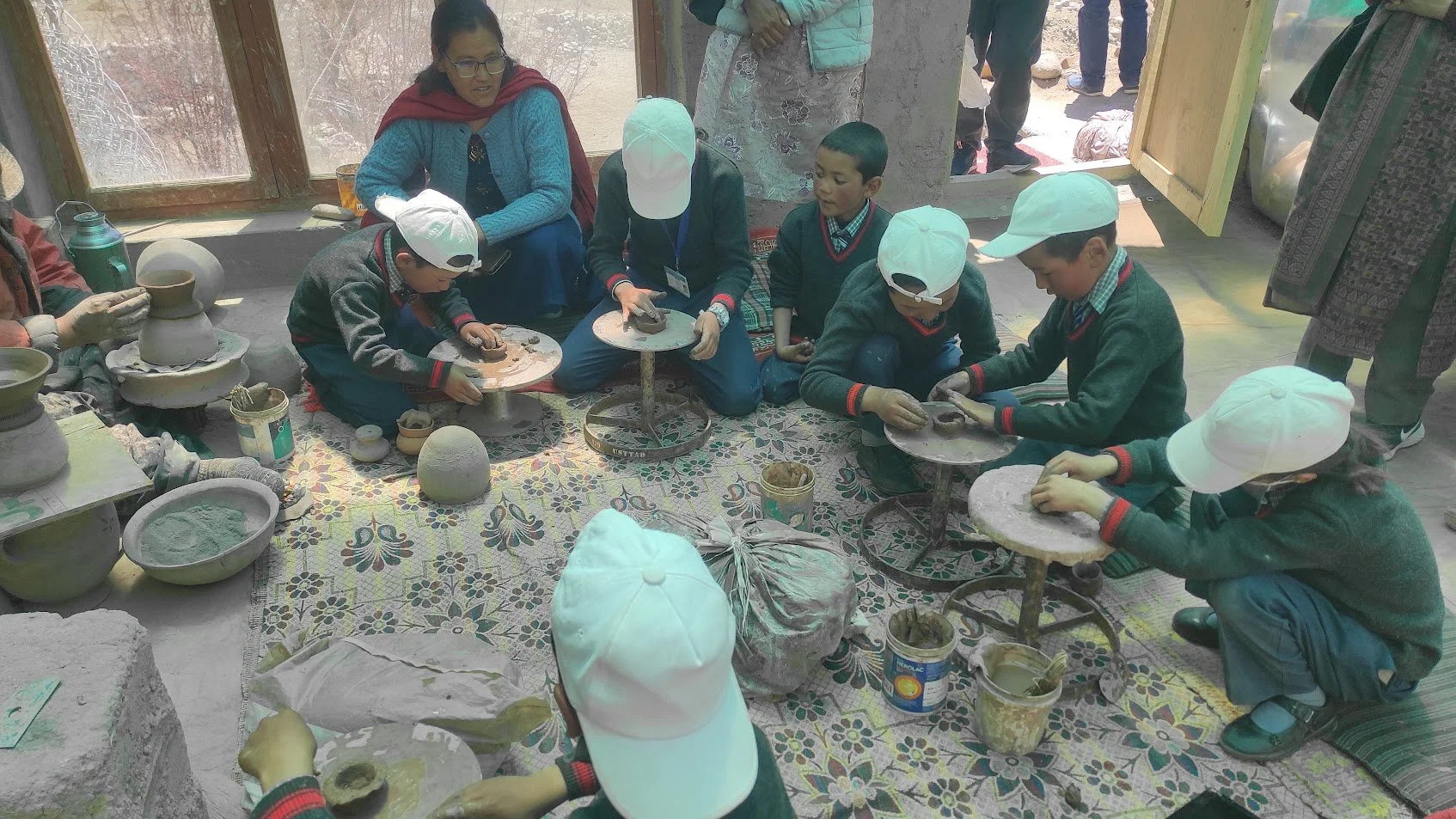 Children engaged in pottery making with adult supervision in a workshop. They are working on small clay pots using hand tools, with various pottery supplies and finished pieces nearby. The setting appears to be indoors with natural light coming throu