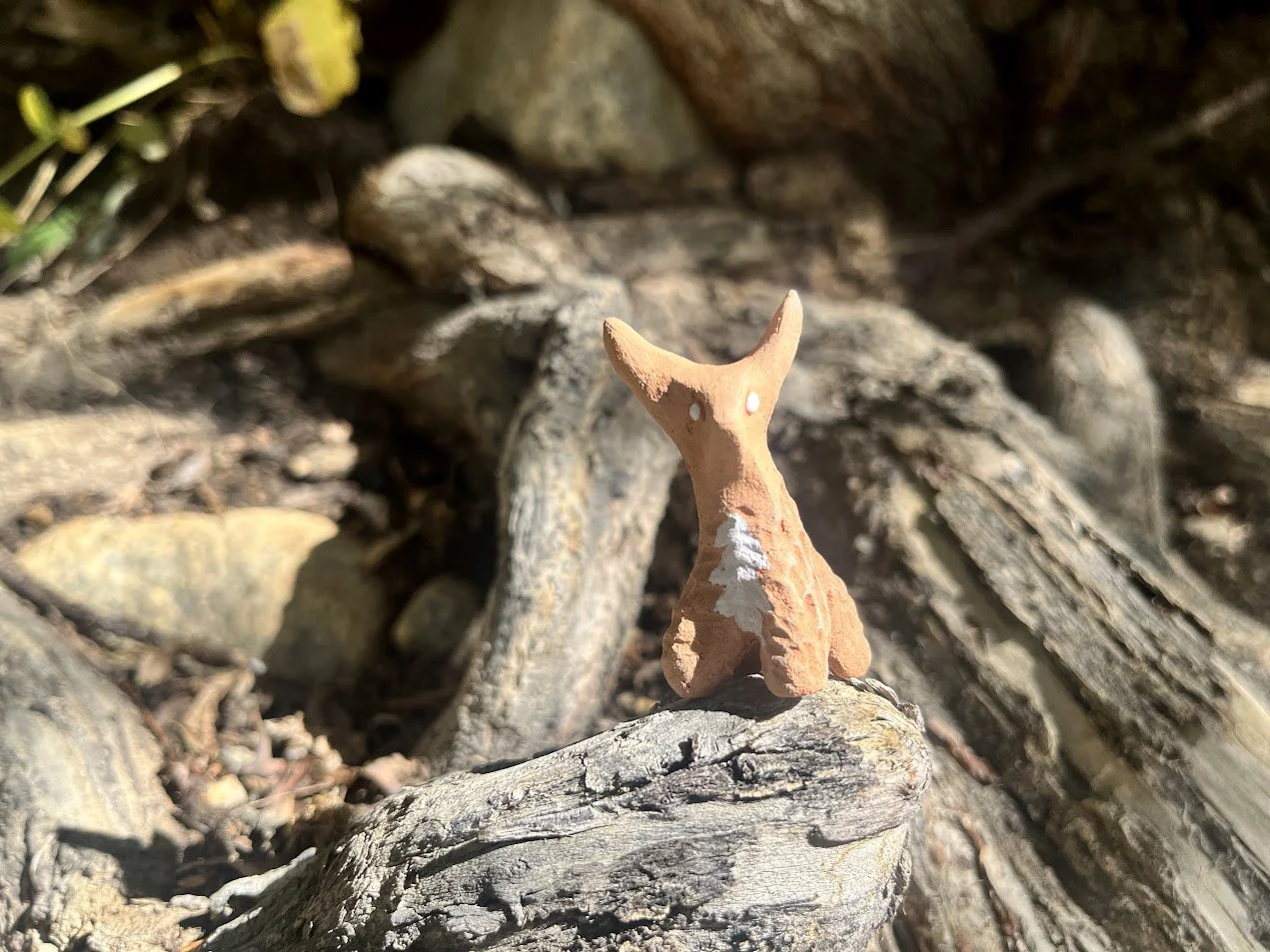 Small terracotta sculpture of a dog with pointed ears, placed on a piece of weathered wood among natural outdoor debris.