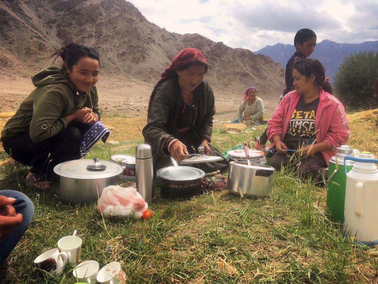 Group of women and children sitting on grass outdoors, preparing and serving food with cooking pots and utensils, in a mountainous landscape with cloudy sky.