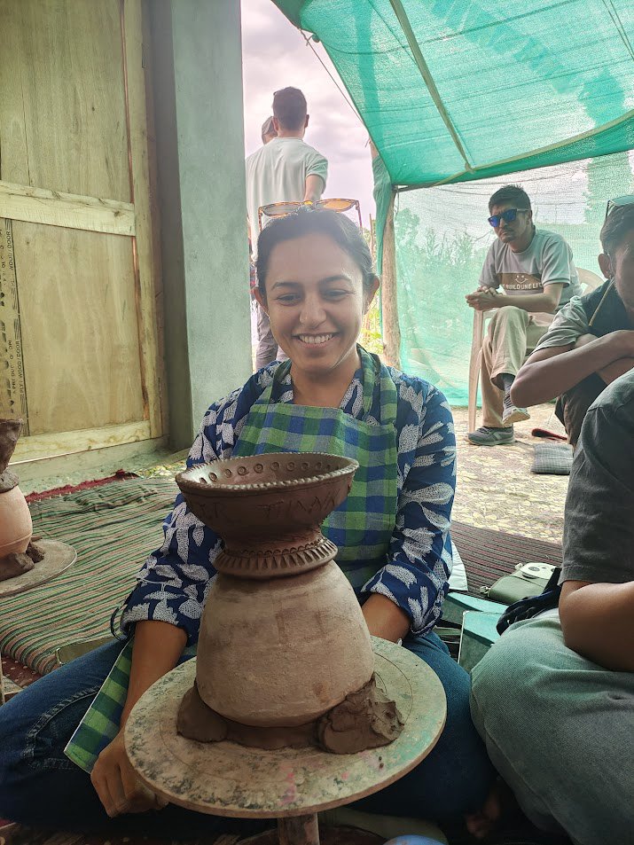 Smiling woman in a blue and green checkered scarf sitting behind a pottery wheel with a partially shaped clay bowl, indoors with other people around.