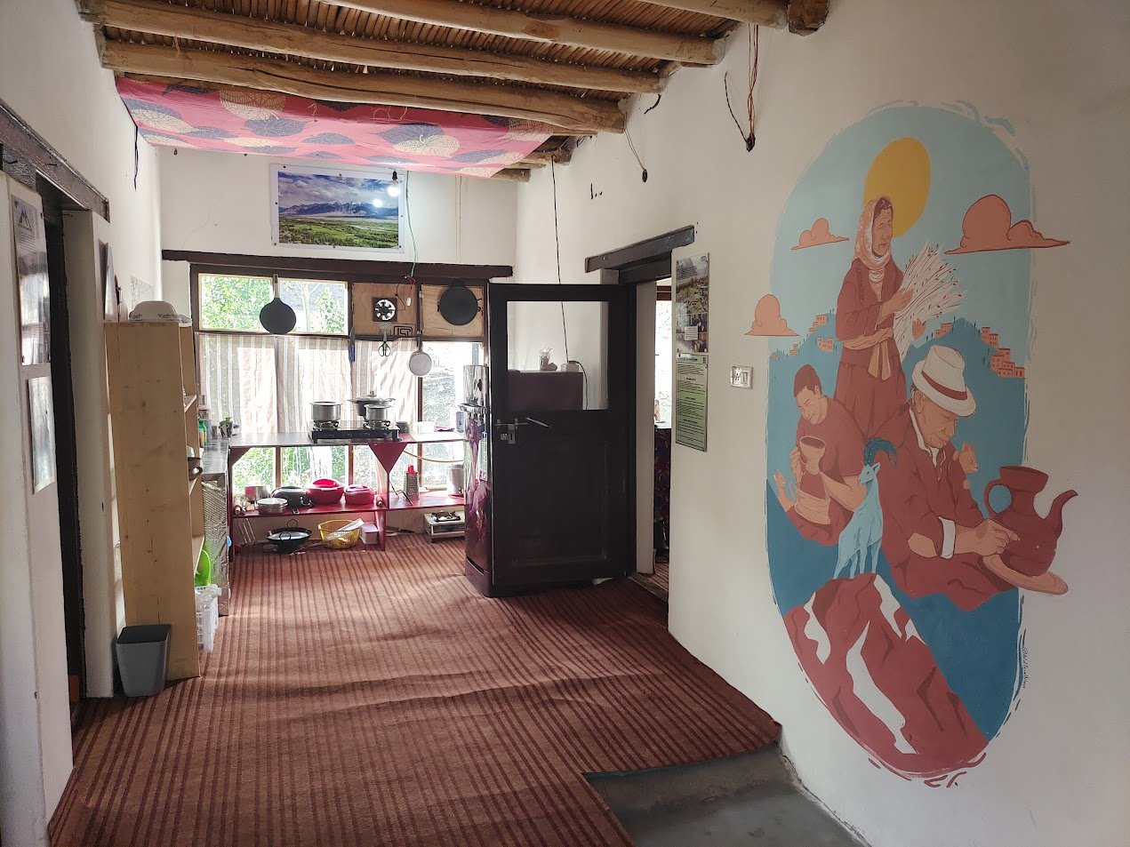 Interior of a rustic kitchen with a wooden ceiling, colorful mural of a woman and two men on the right wall, and a window with outdoor scenery in the background.