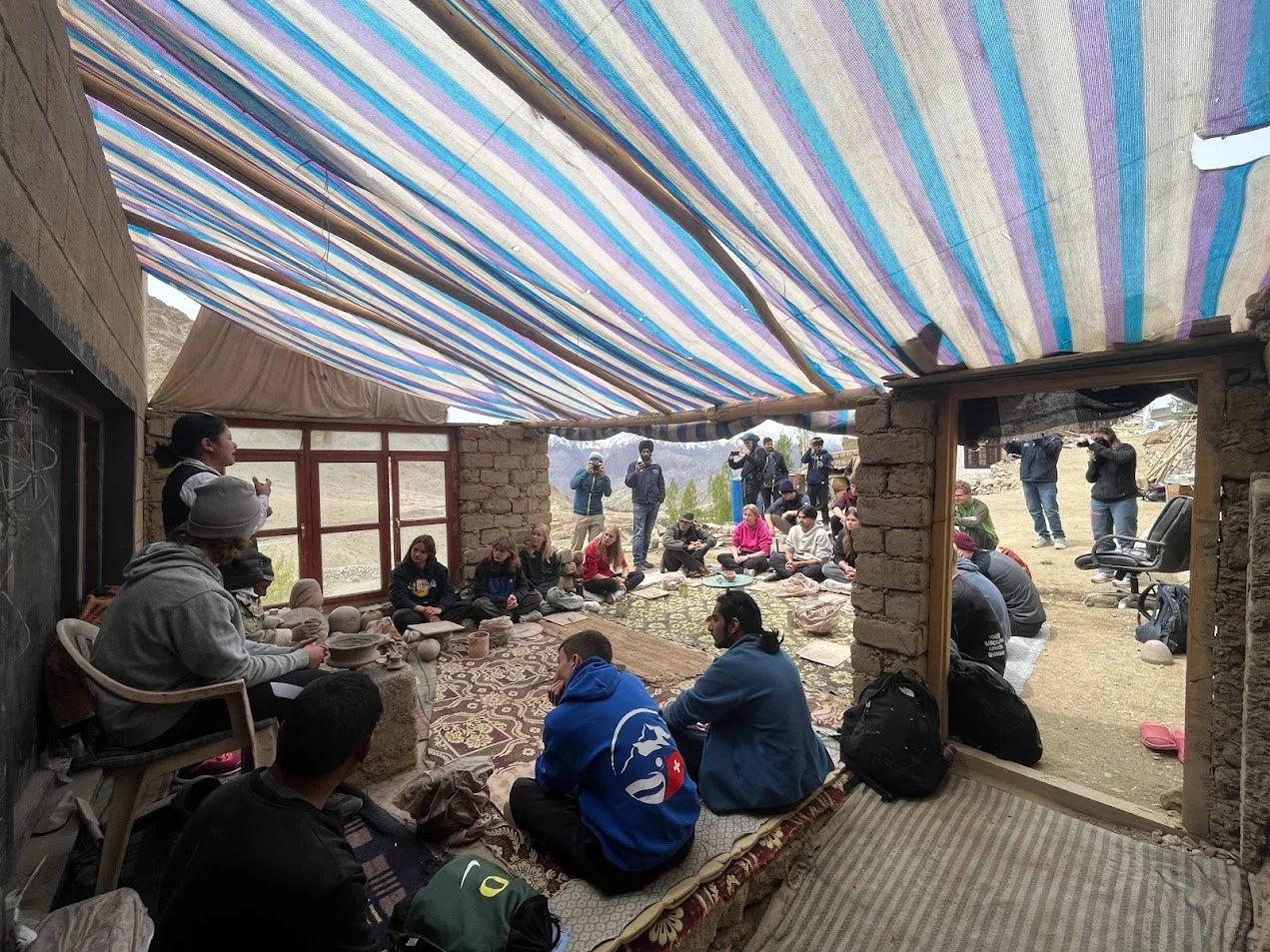 Group of people sitting inside a partially open room with a makeshift roof made of striped fabric, some standing outside, in a scenic outdoor setting.