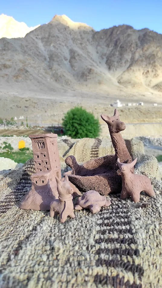 Small clay animal figurines, including a horse, cow, and other animals, arranged outdoors on a textured fabric with mountain landscape in the background.