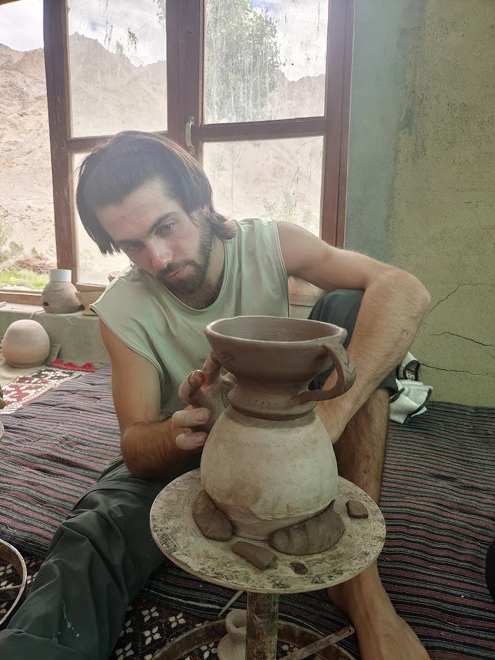 A man shaping a clay pot on a pottery wheel inside a room with natural light coming through the window.