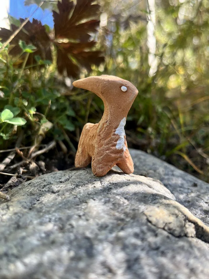 Small clay sculpture of a creature with a beak-like nose, white eyes, and a white patch on the chest, placed on a rock outdoors surrounded by green foliage.