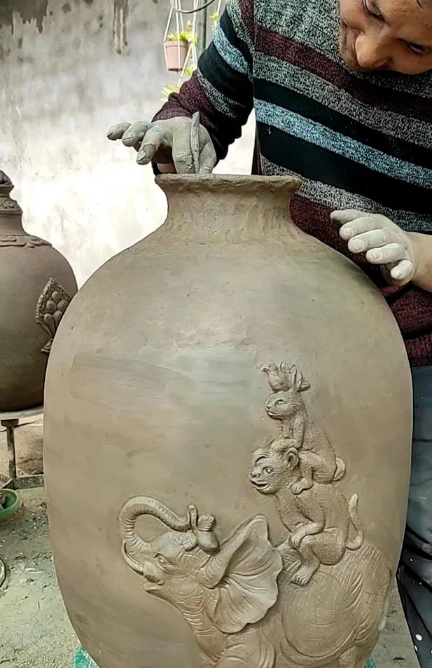 A man creating a large clay vase with detailed elephant and monkey carvings on its surface.