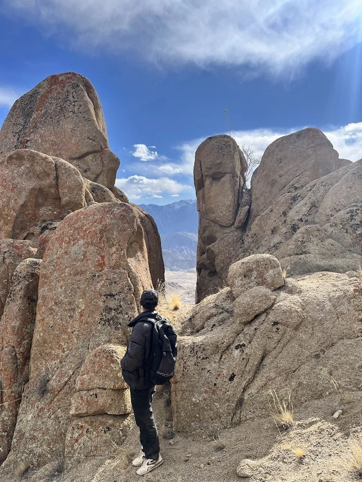 A person wearing a black jacket and backpack standing among large desert rocks with mountains and blue sky in the background