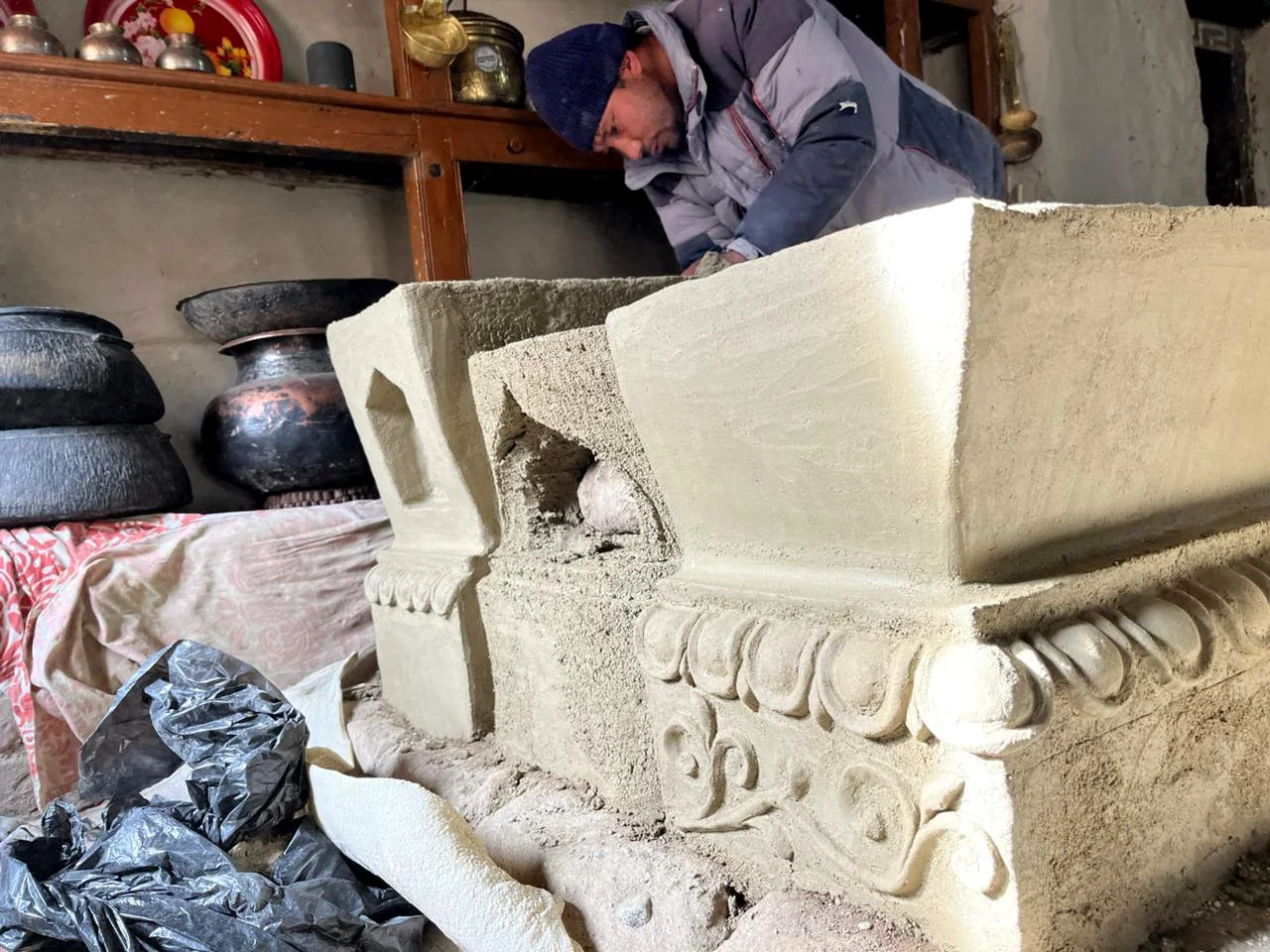A man dressed in a jacket and knit cap working on carving or restoring a large clay or architectural element inside a workshop or similar space.