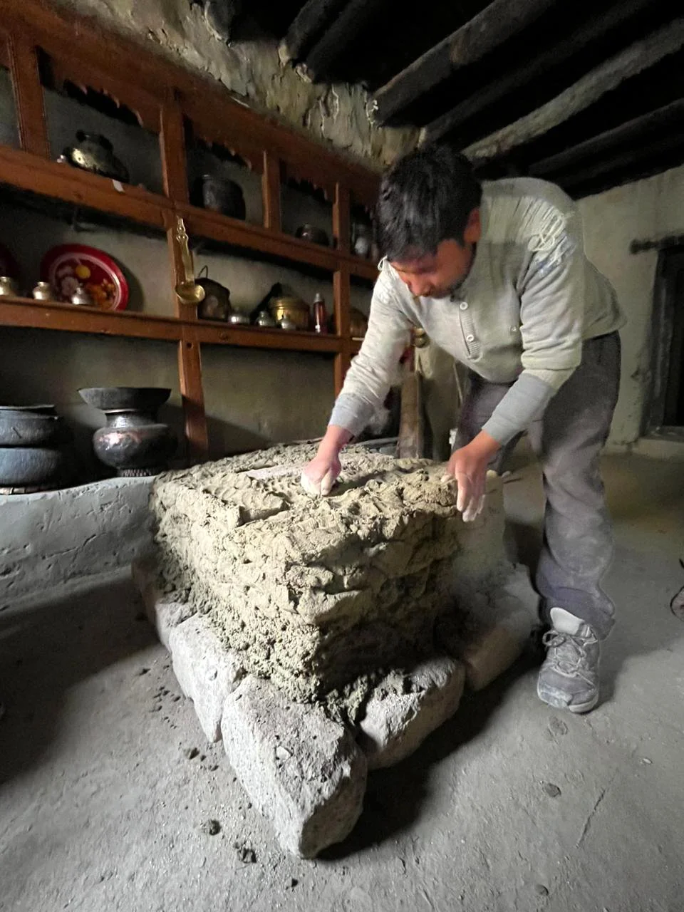 A man working on a mud brick in a rustic room with shelves of pottery and metalware on the wall.