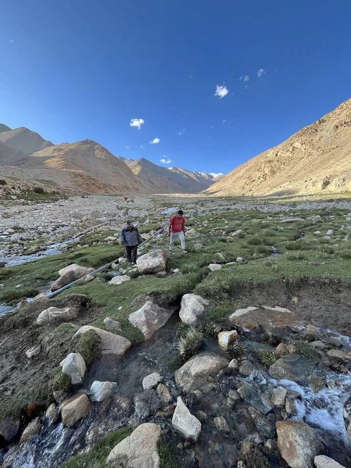 Two people standing on a grassy, rocky mountain valley with a small stream, surrounded by high mountains under a clear blue sky.