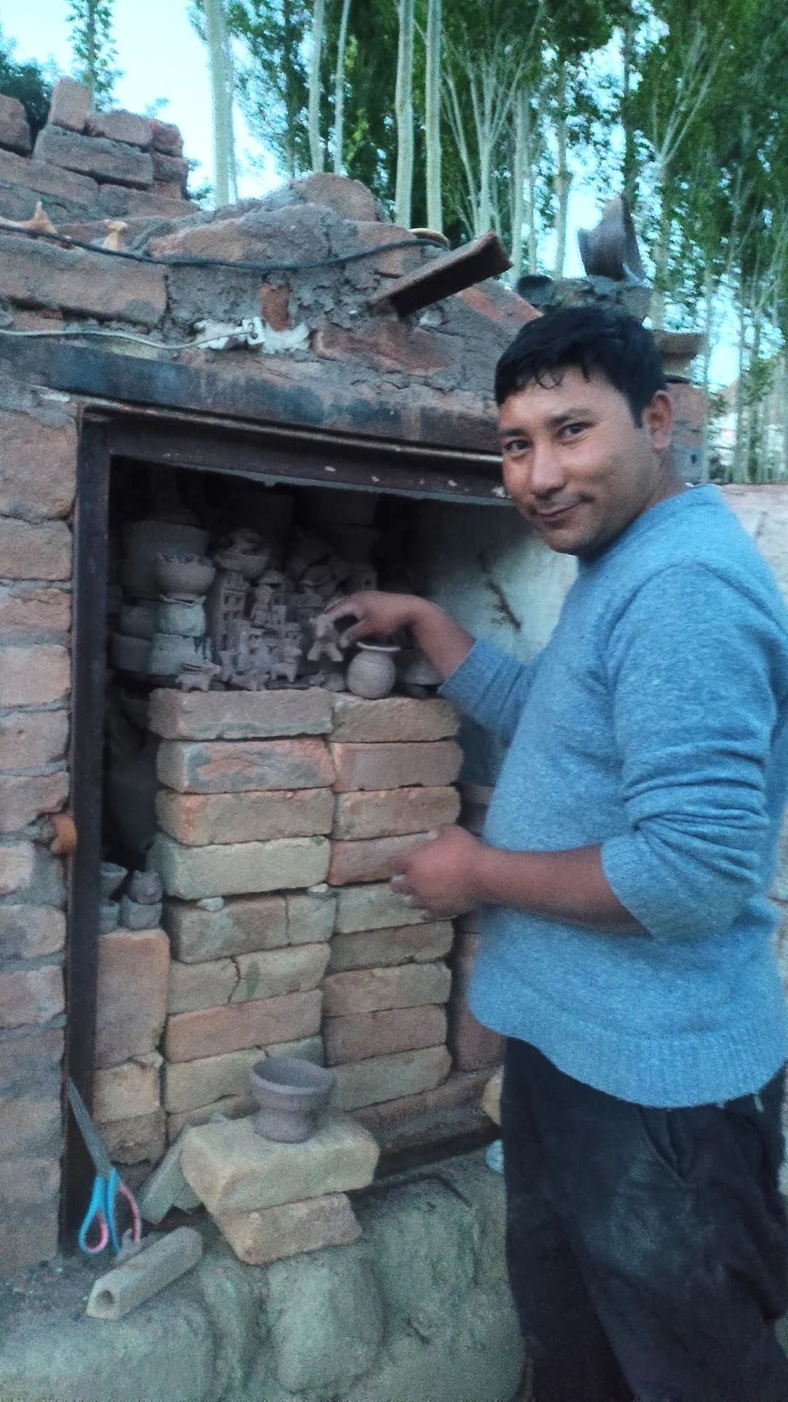 A man wearing a gray shirt stands next to a brick kiln, reaching towards the inside. The oven is built with bricks and has a metal door, with various small clay pots and figurines inside. There are green trees in the background.
