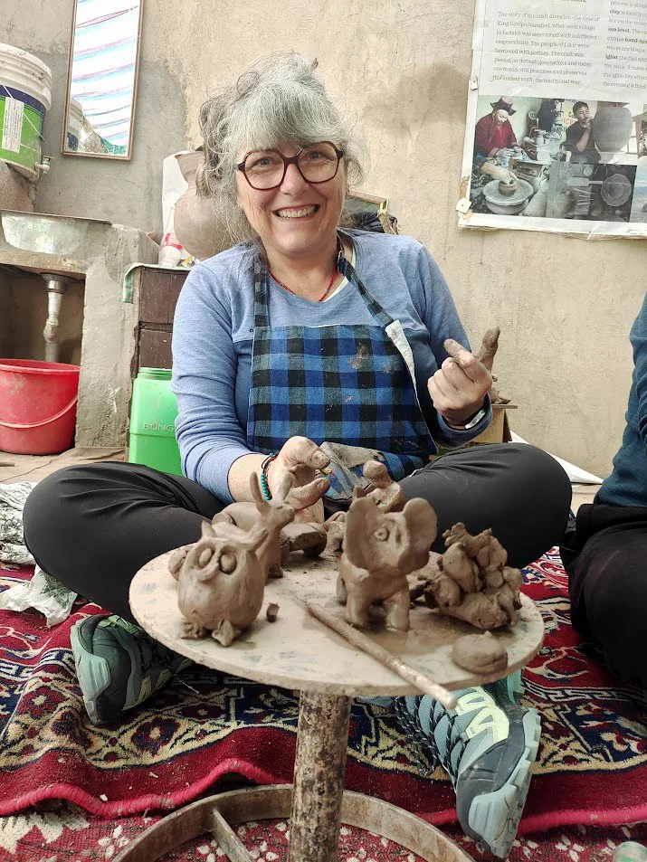 A woman sitting cross-legged on a colorful rug, smiling while making clay sculptures of mice. She wears glasses, a blue shirt, and a checkered apron. The background includes a textured wall with a poster and some tools, with various objects around.
