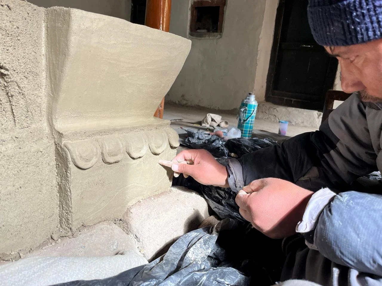 A person wearing a blue beanie and gray shirt working on a decorative clay carving on a building's exterior wall, with various tools and supplies nearby.