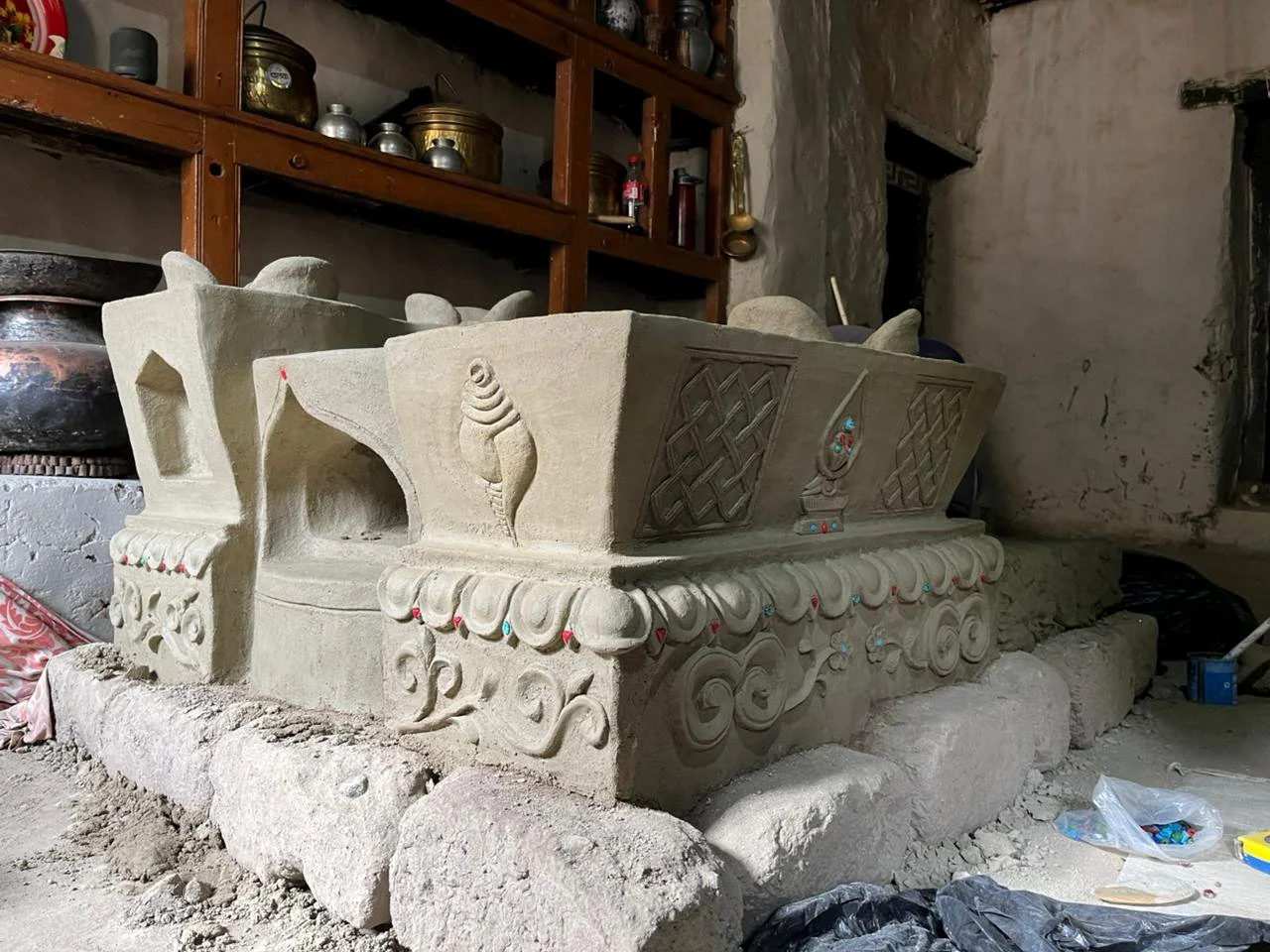 Unfinished clay stove (thap) with carvings and decorations, placed on large rocks, in a room with shelves holding jars and bottles.