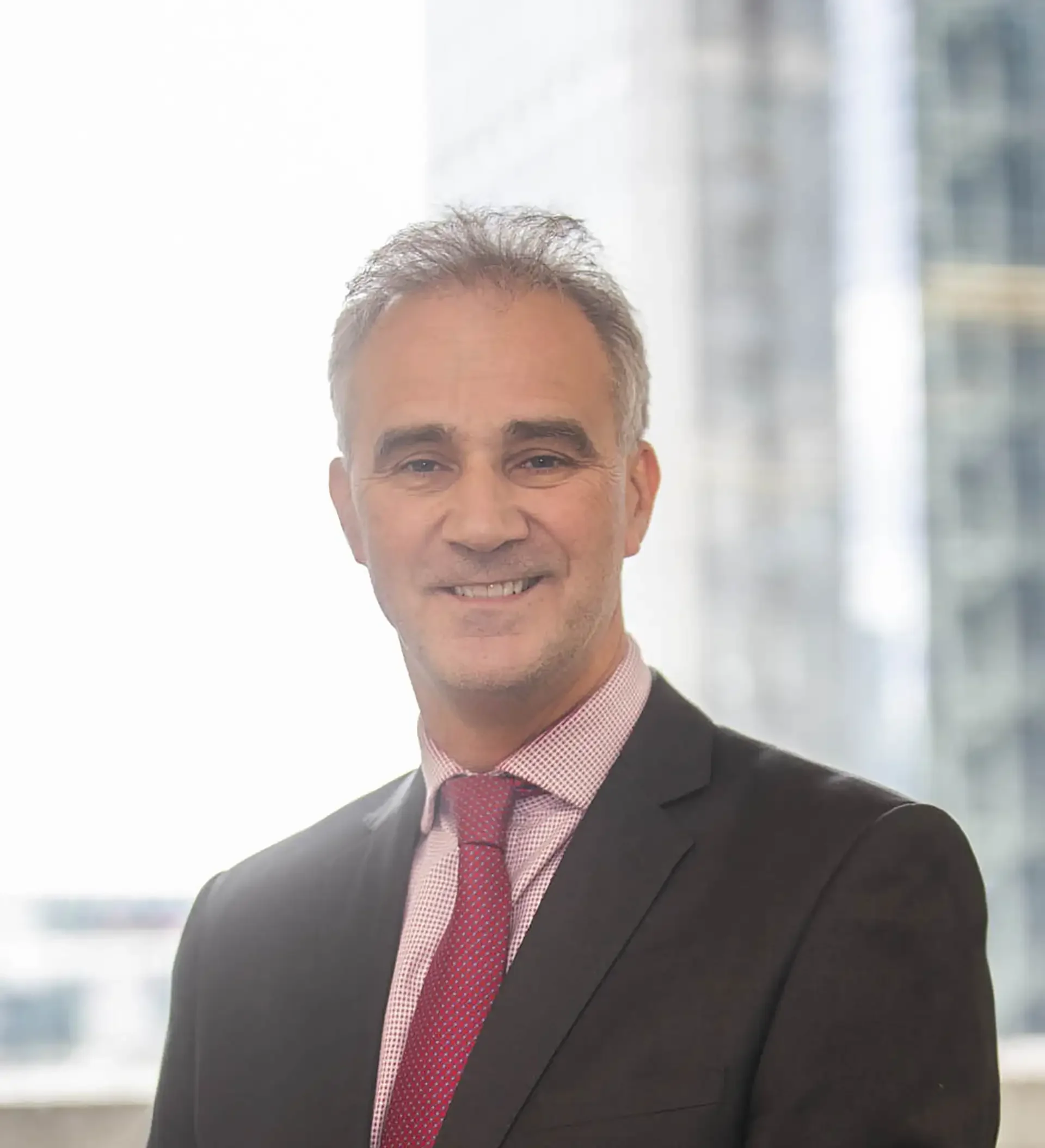 A middle-aged man wearing a business suit, pink dress shirt, and red tie, smiling in a professional setting with a blurred office background.