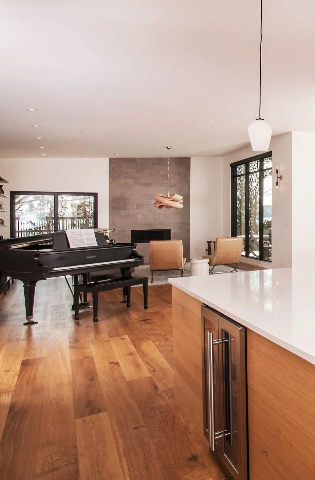 Modern living room with a black grand piano, beige armchairs, a stone fireplace, large windows, wooden flooring, and a white kitchen island.