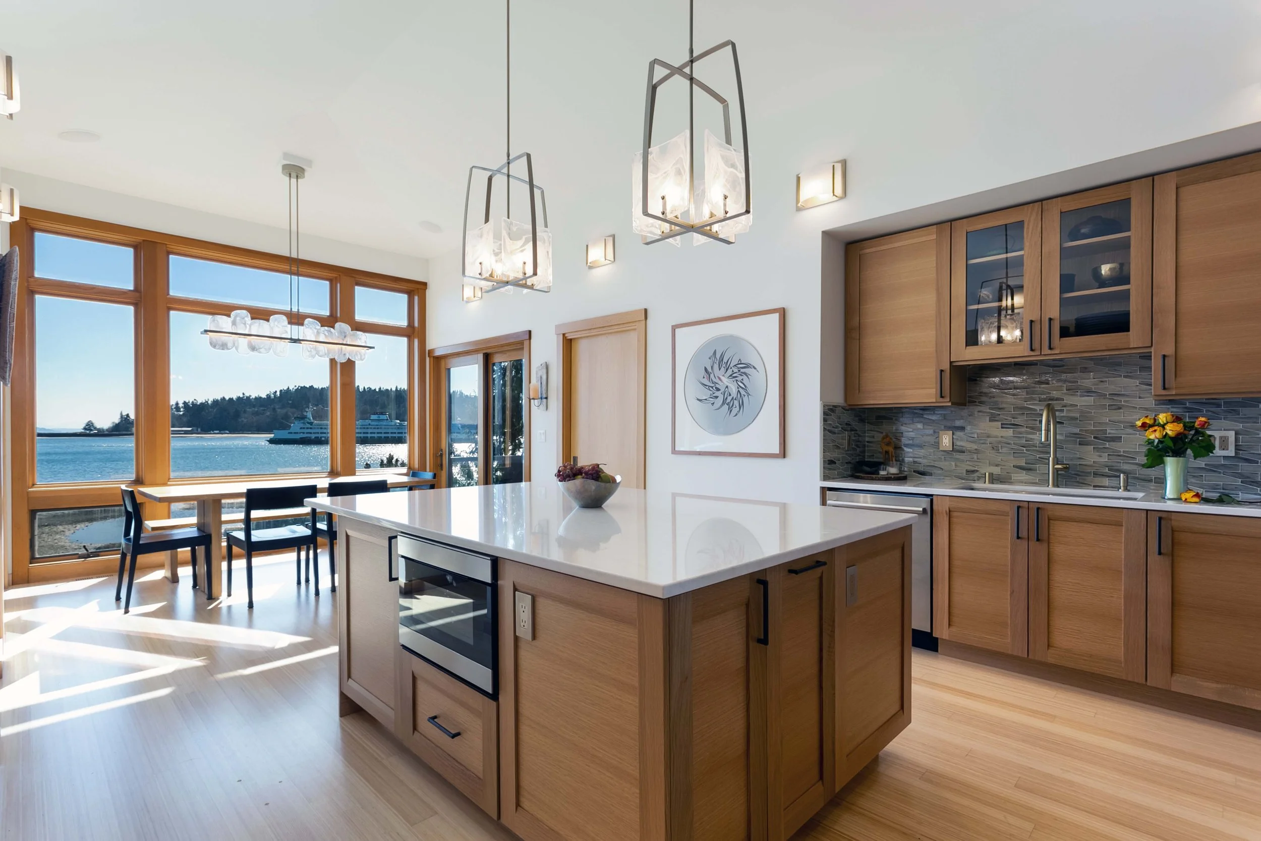 A modern kitchen with wooden cabinets, a white island with a built-in oven, and large windows overlooking a body of water. There are hanging pendant lights above the island and a dining table with chairs nearby. | McCabe By Design