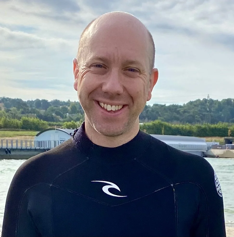 Nigel Lane smiling wearing a black wetsuit outdoors near water, with a background of trees, hills, and a cloudy sky.