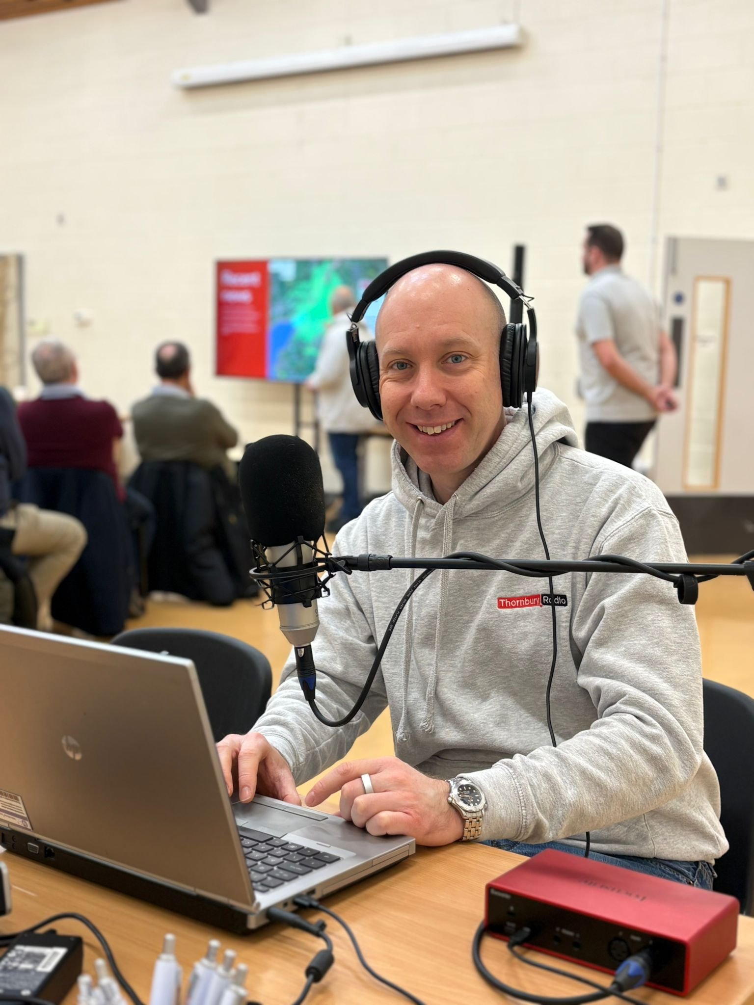 Nigel Lane wearing headphones and a light gray hoodie seated at a table with a laptop and microphone, in a room with several people and a large screen in the background.