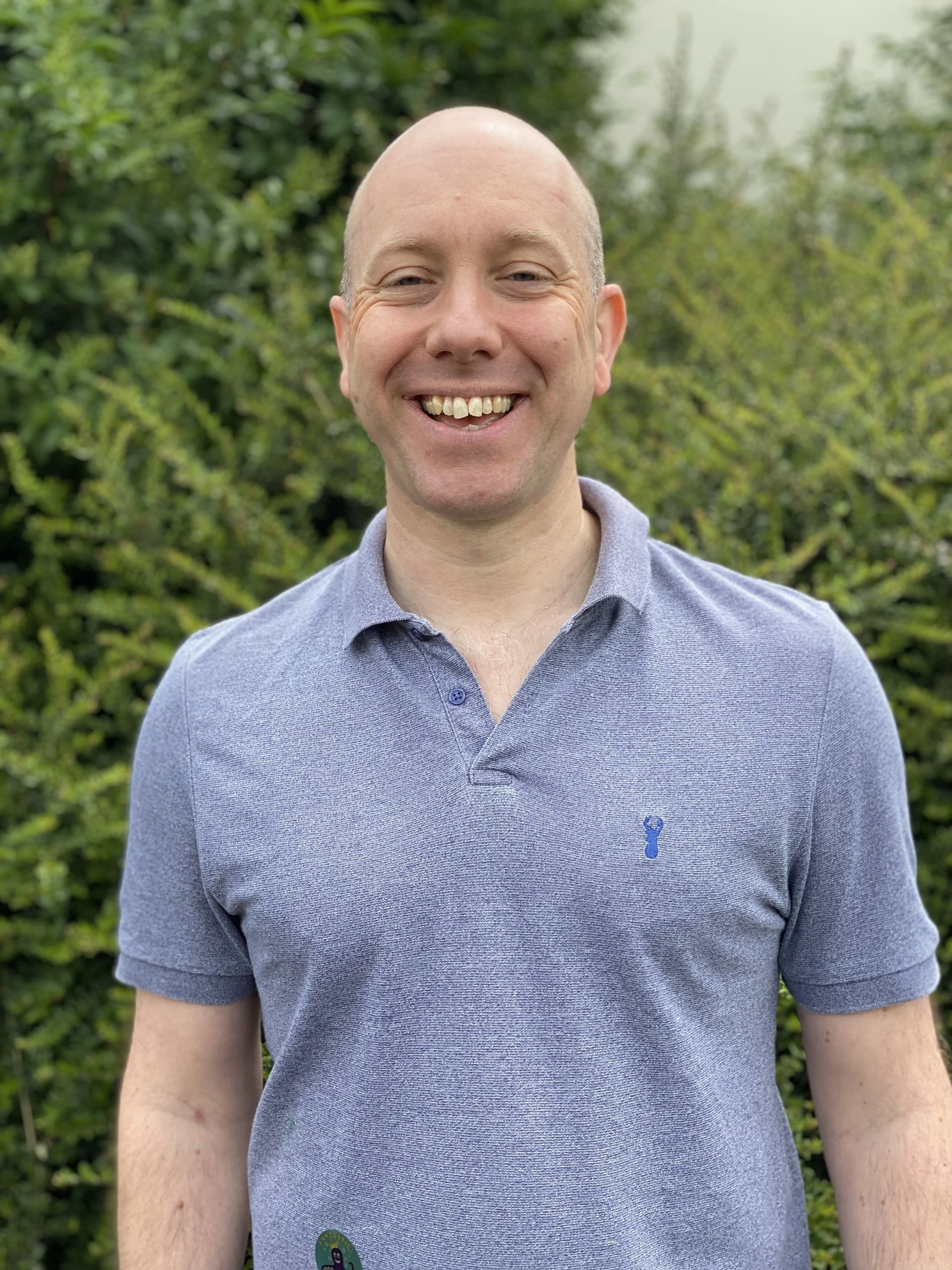 Nigel Lane smiling, wearing a blue polo shirt, standing outdoors with green foliage in the background.