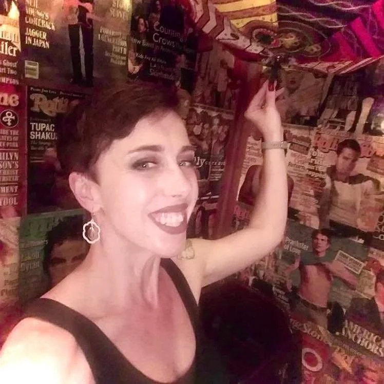 Amanda Lang with short dark hair smiling and wearing earrings, raising her arm in a room decorated with numerous posters and magazine covers on the walls.