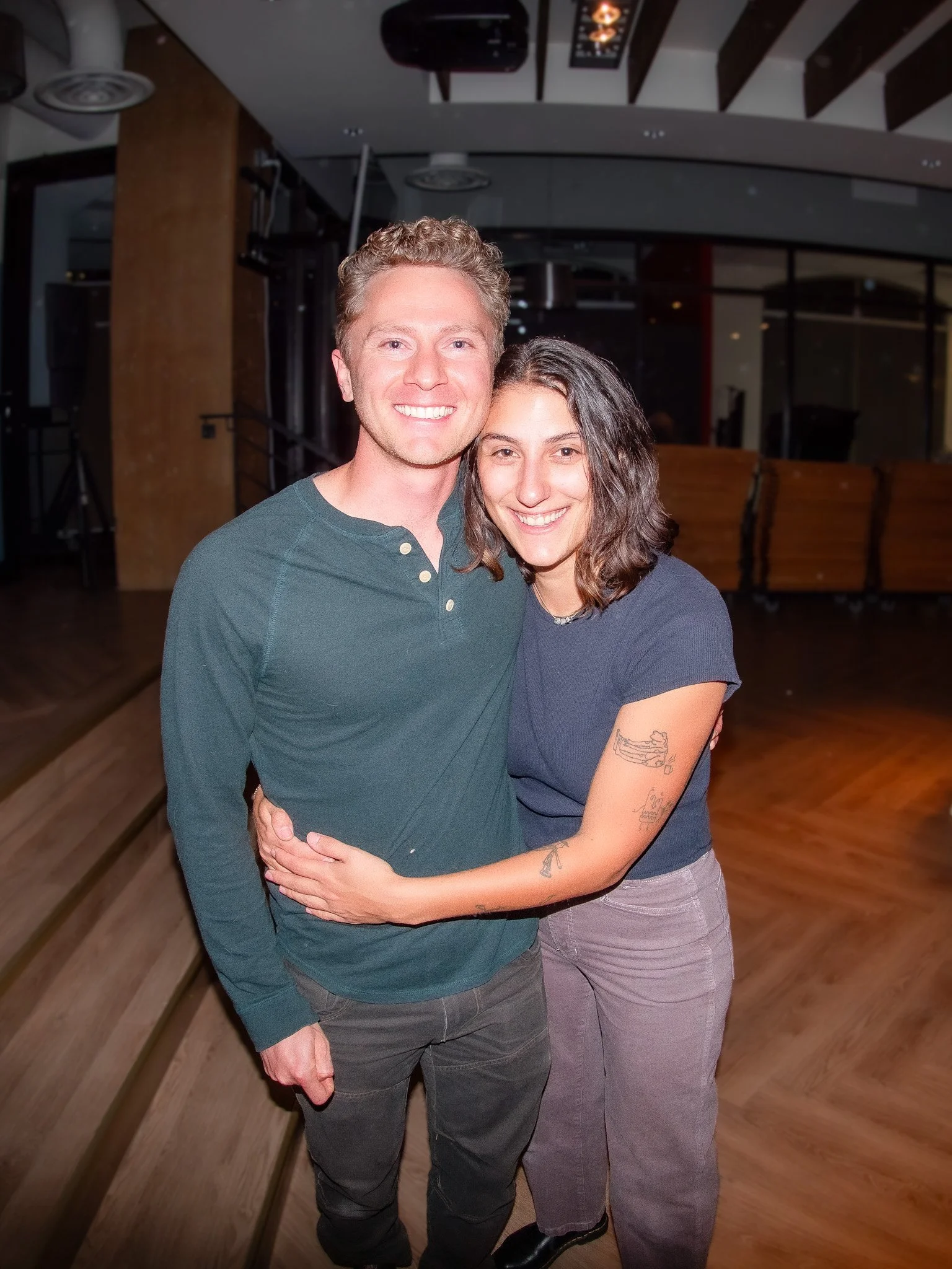 A man and woman smiling and hugging indoors, with a wooden floor and dim lighting.