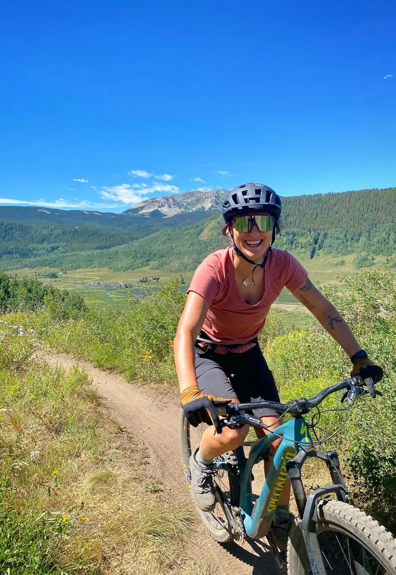 Woman mountain biking on a dirt trail in a scenic mountainous area with green hills, trees, and a clear blue sky.