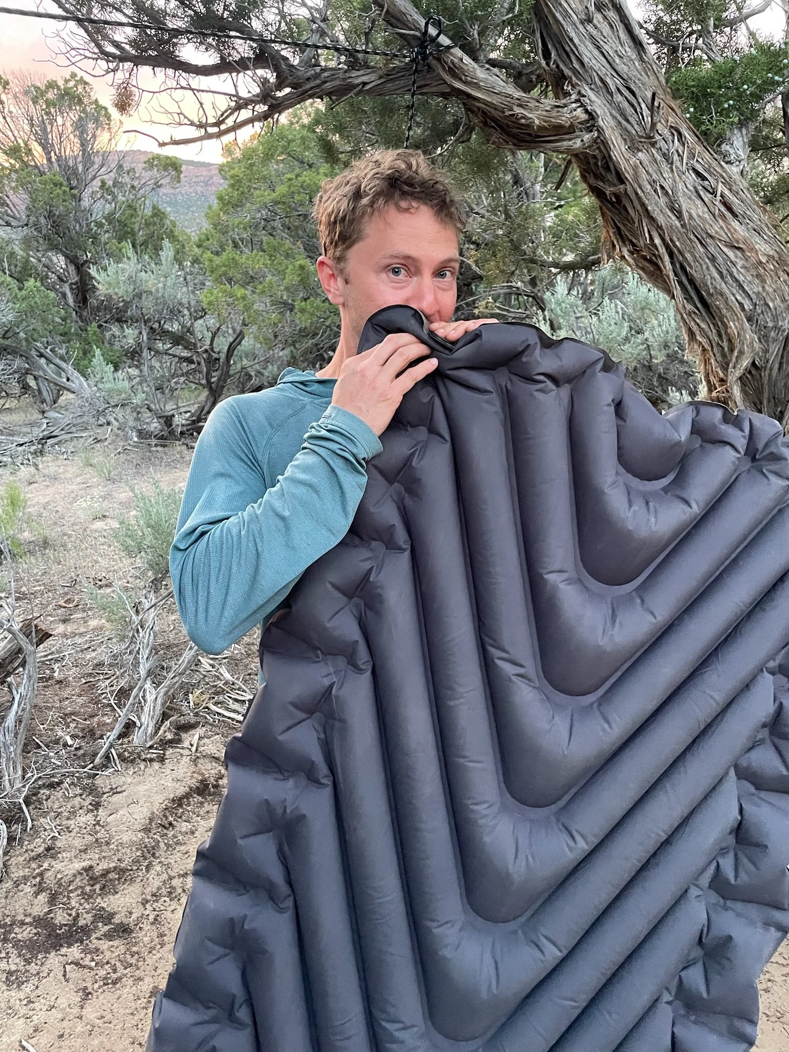 Person standing outdoors in a dry, wooded area holding a large black inflatable sleeping pad, with twisted tree branches overhead and green shrubs in the background.