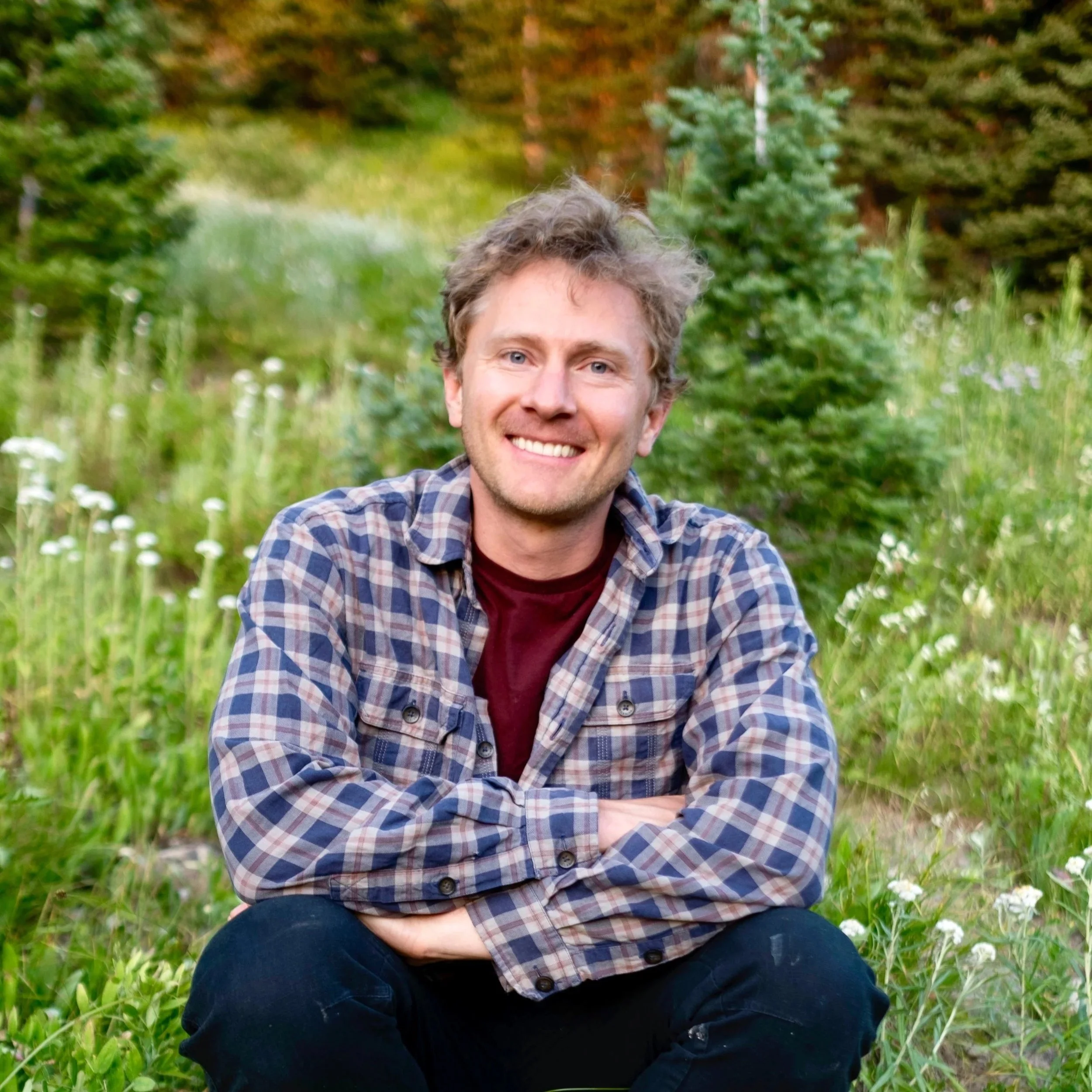 Jacob, co-founder of GoodPair Studio, seated outdoors in a mountain meadow surrounded by wildflowers and evergreen trees