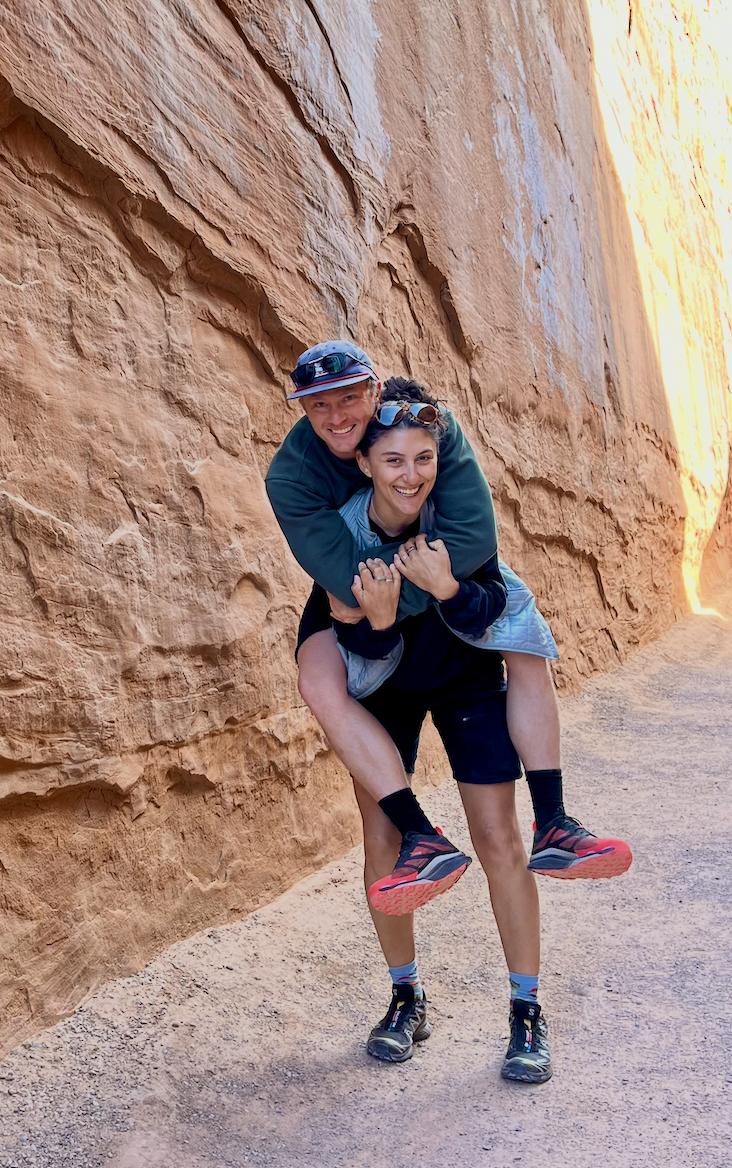 Two hikers, a man and a woman, enjoying an outdoor adventure with a rock formation in the background. The woman is giving the man a piggyback ride, both smiling.