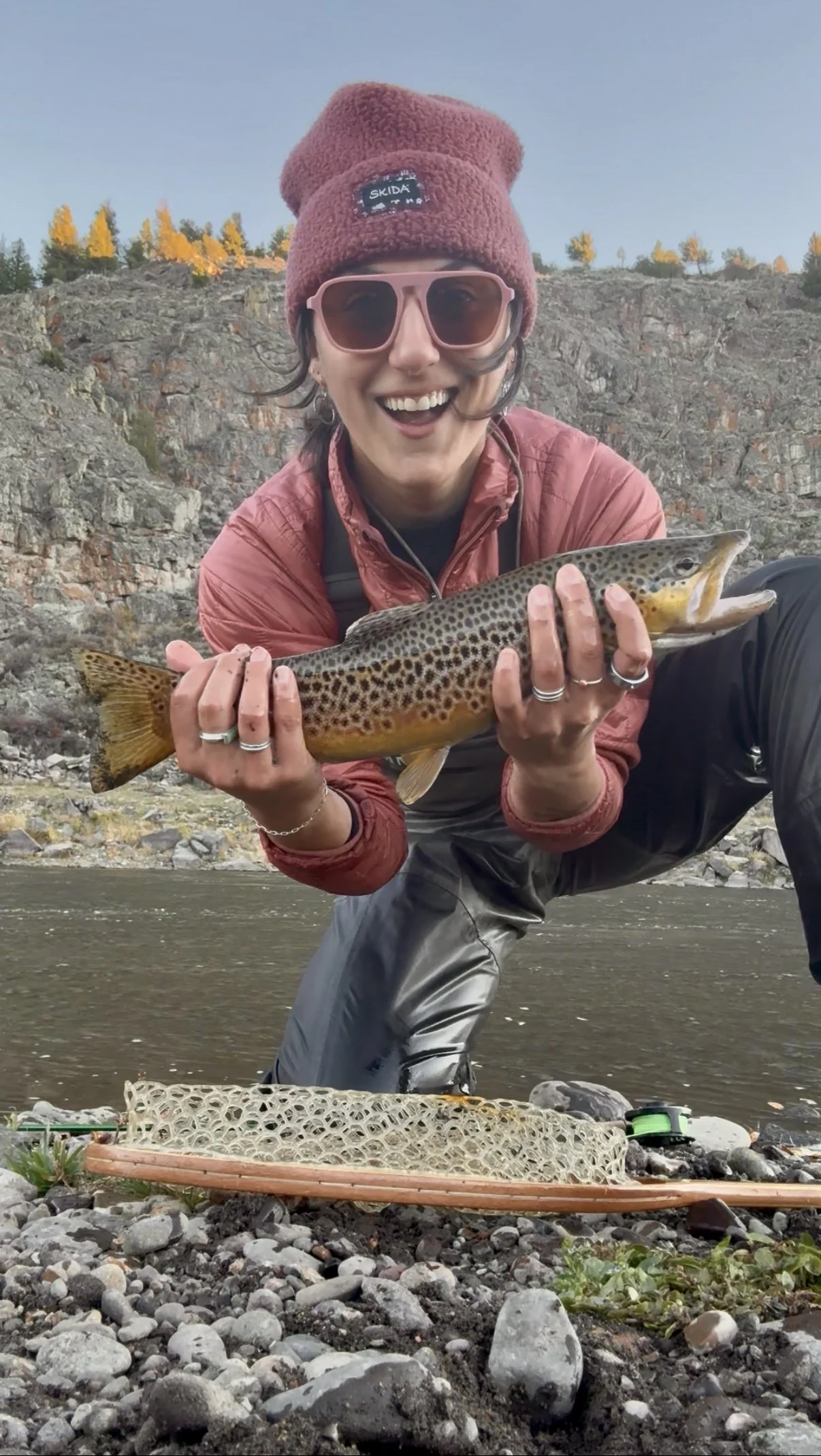 Person wearing a pink beanie, pink sunglasses, and a red jacket holding a large fish while kneeling on rocks near a river with hills in the background.