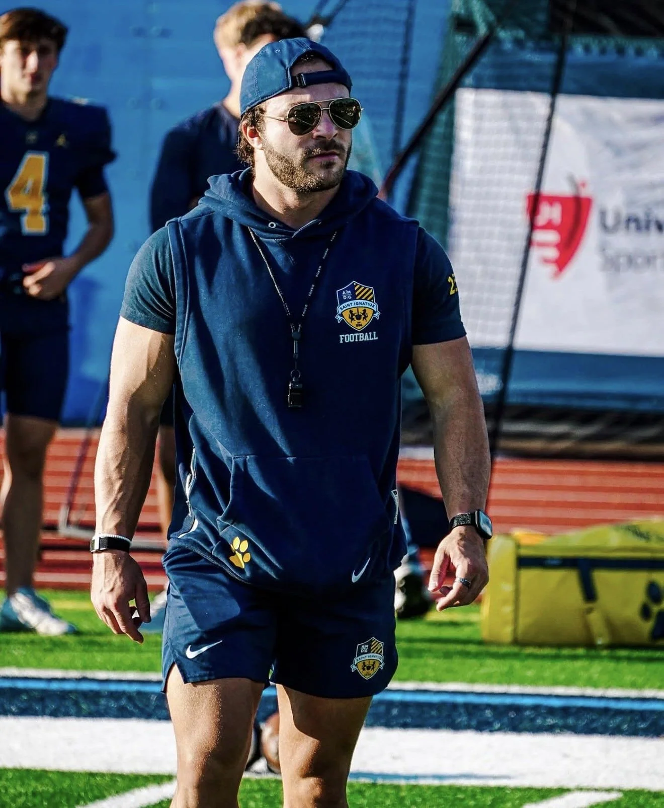A man wearing sunglasses, a blue cap backward, and sportswear with a football team's logo, stands on a sports field.