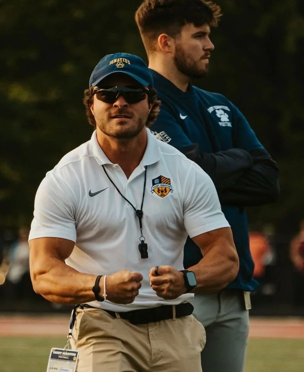 Two men on a sports field, one wearing sunglasses, a cap, and a white polo shirt with a logo, and the other in a blue sports jacket with arms crossed, in a scene with trees in the background.