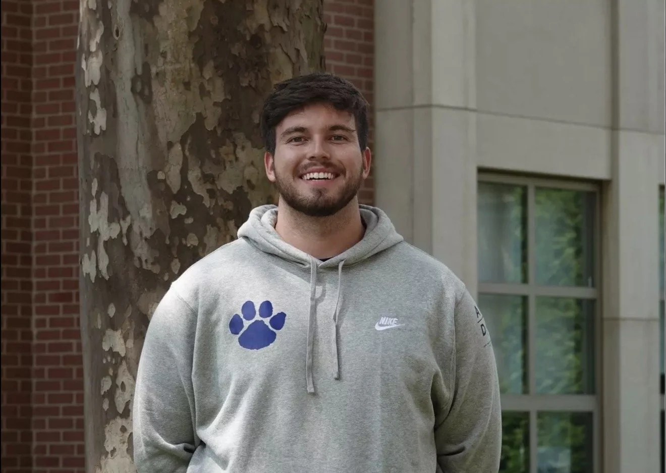 Young man smiling outdoors near a tree and modern building, wearing a gray hoodie with a blue paw print and Nike logo.